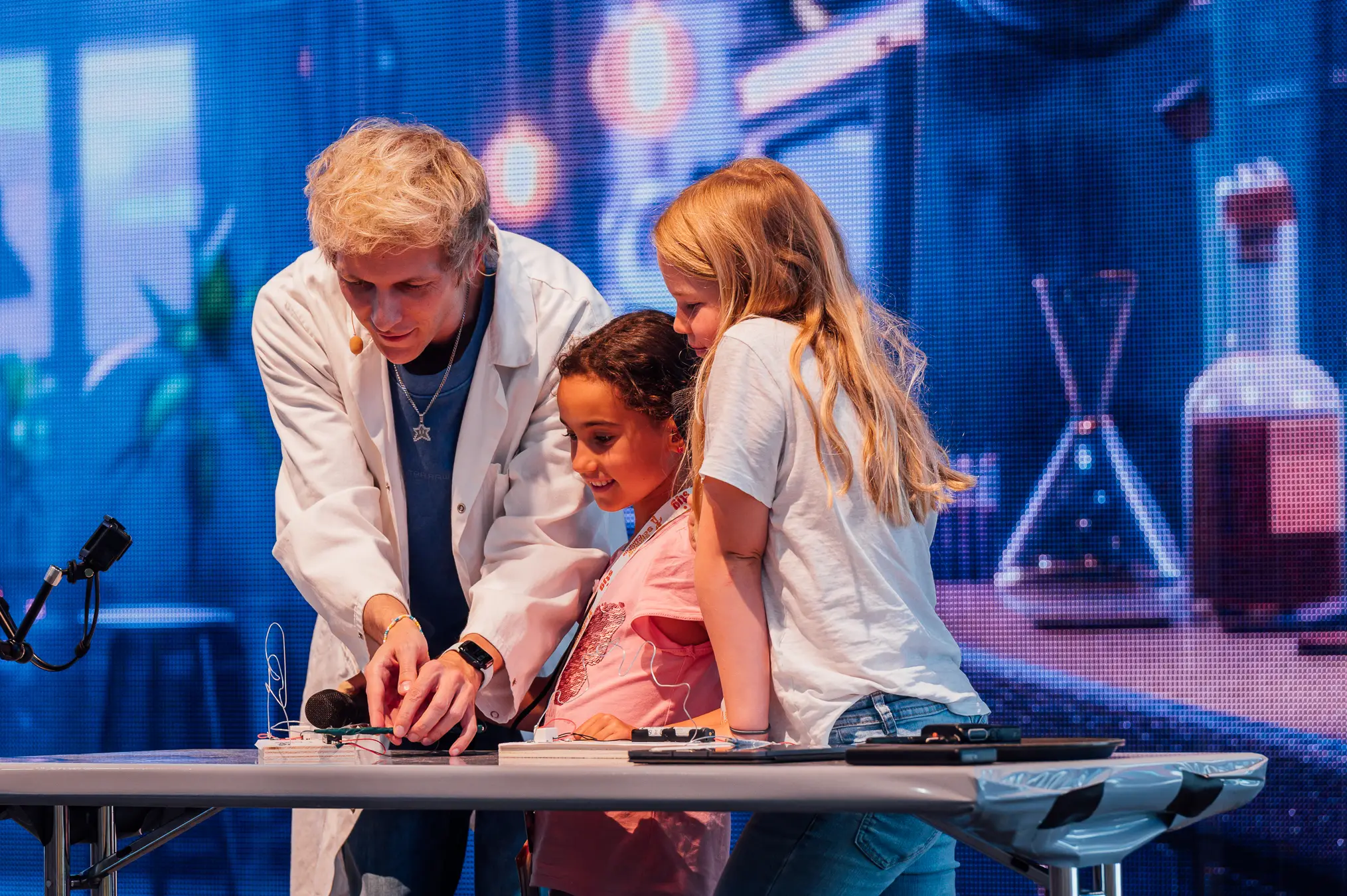 Mitarbeiter im Laborkittel führt auf der Bühne mit zwei Kindern ein Experiment vor