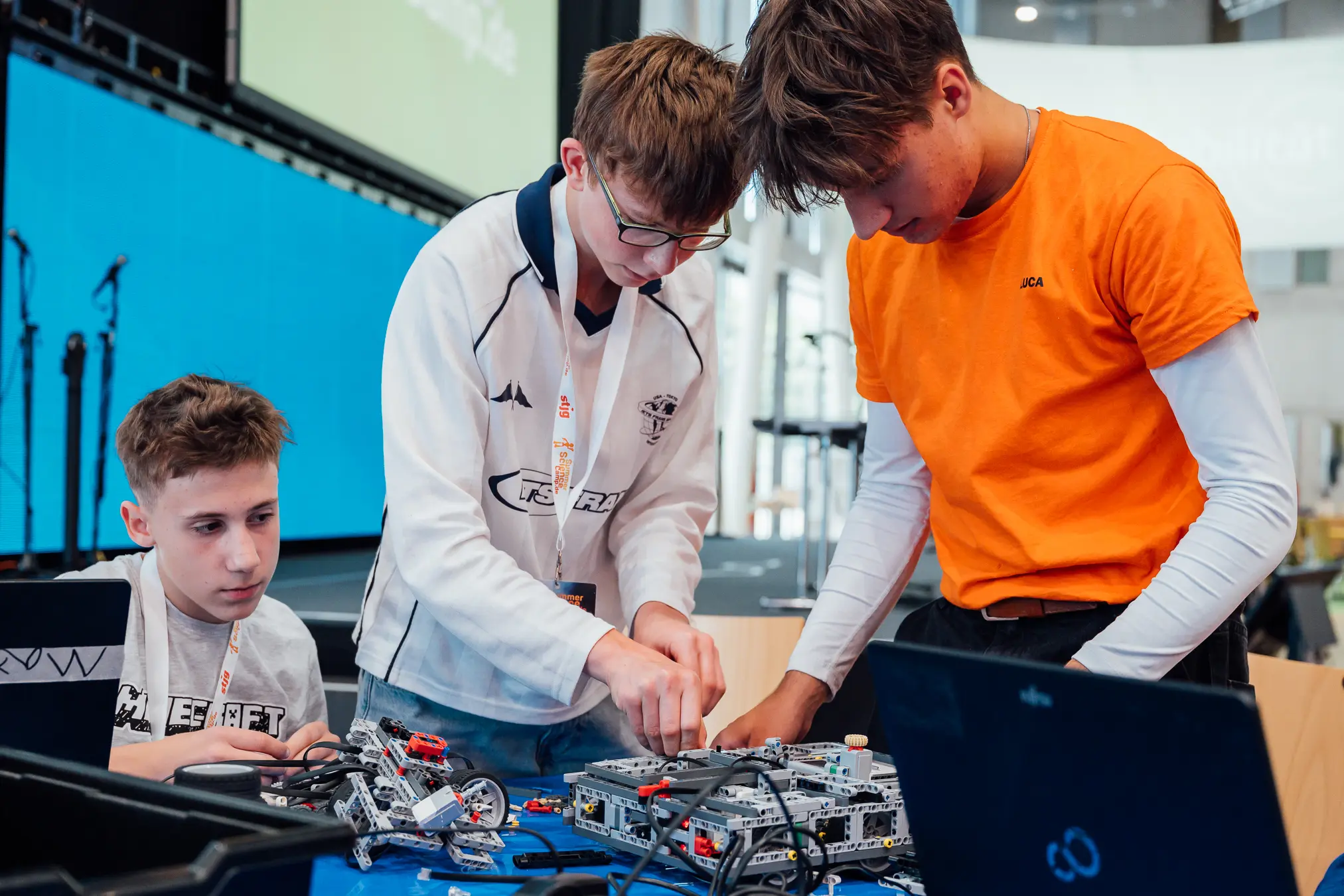 Mitarbeiter im orangefarbenen Shirt hilft zwei Jugendlichen an einem Lego-Roboter, daneben Laptops