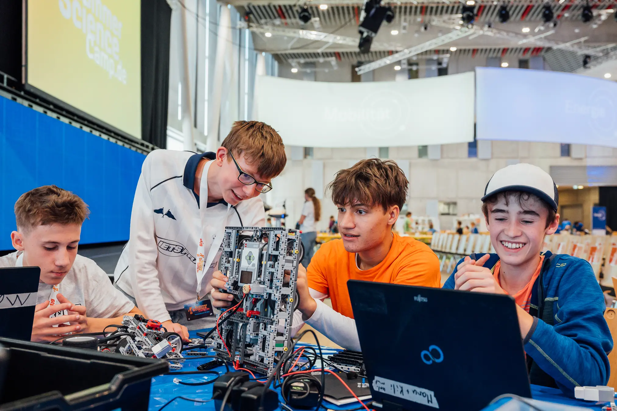 Vier Jugendliche begutachten gemeinsam einen hochgehaltenen Lego-Roboter, davor Laptops