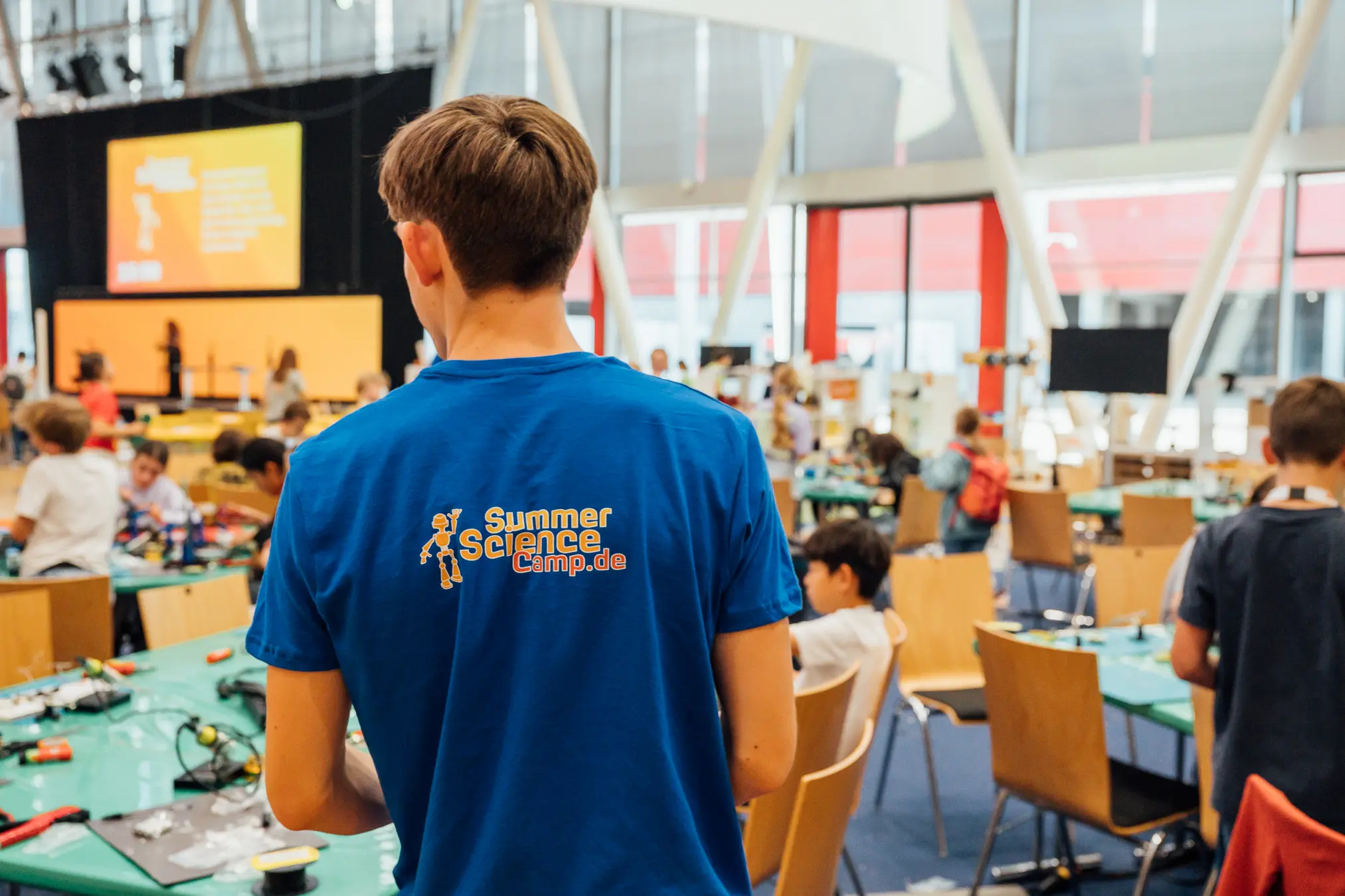 Mitarbeiter von hinten im blauen Shirt mit Summer-Science-Camp-Logo, davor die volle Camp-Halle