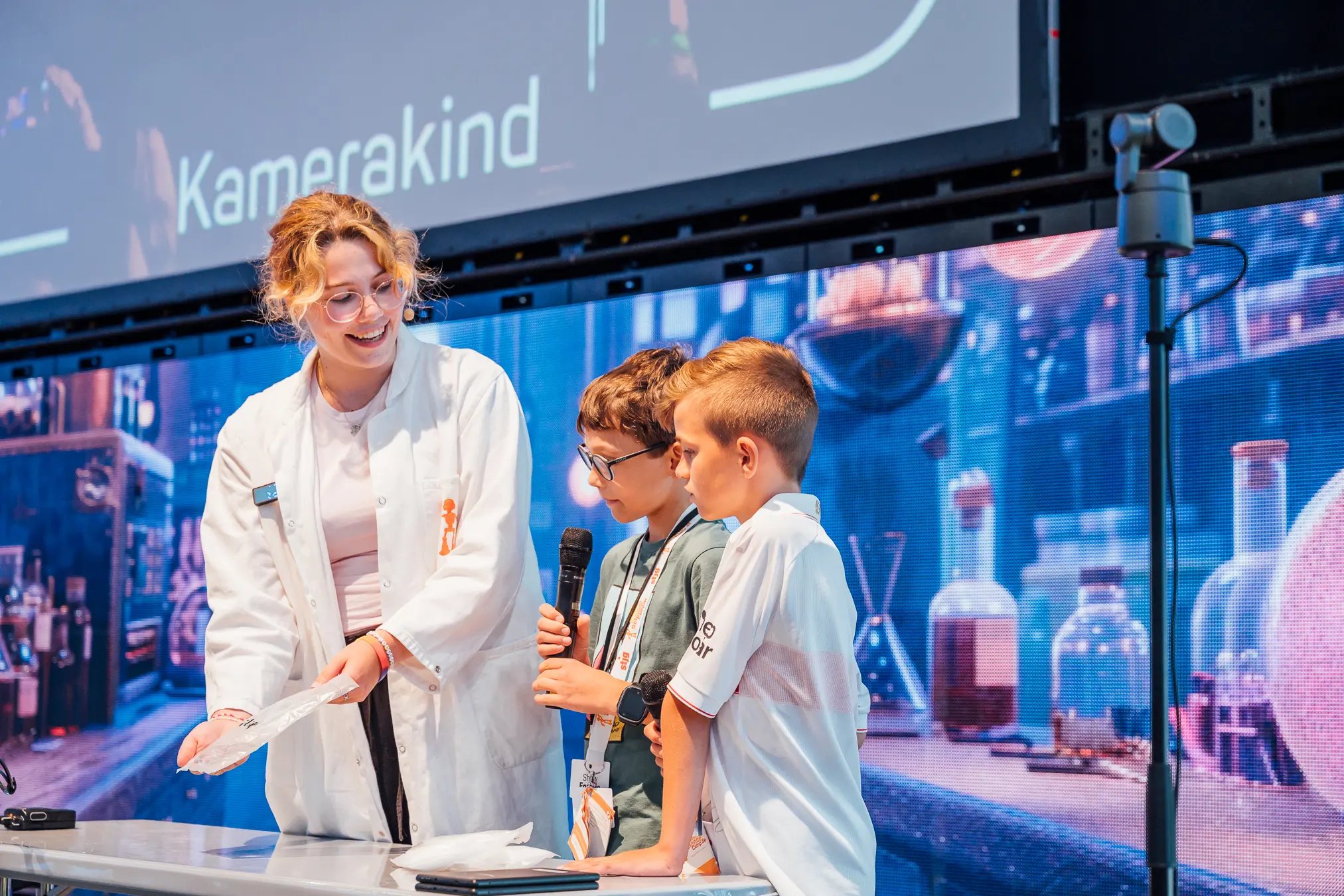 Mitarbeiterin im Laborkittel moderiert auf der Bühne ein Experiment mit zwei Jungen am Mikrofon
