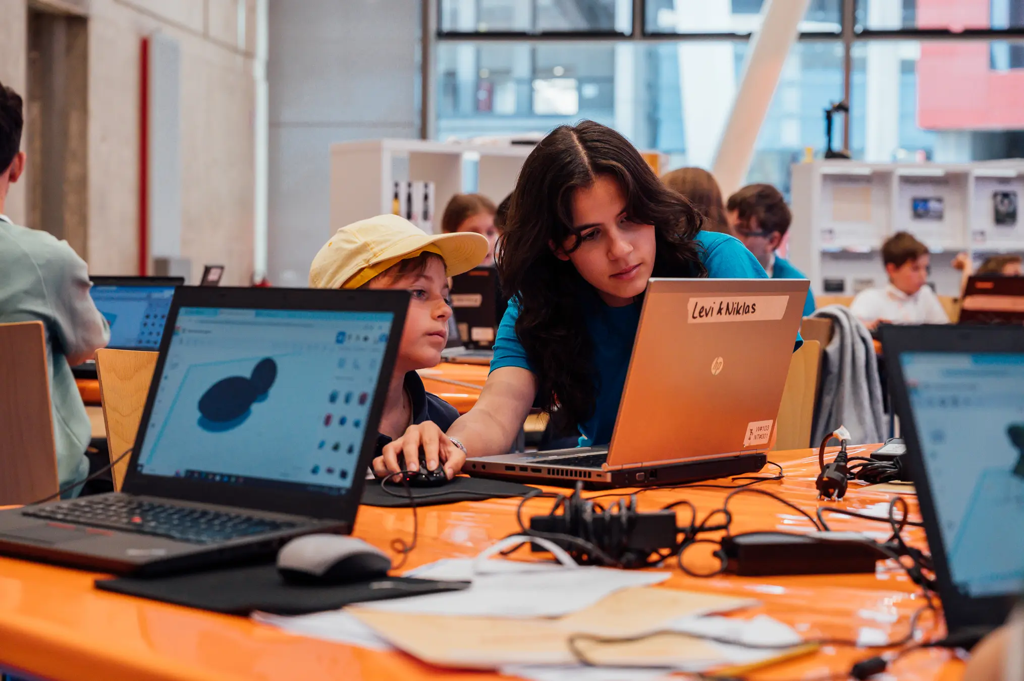 Mitarbeiterin sitzt neben einem Jungen am Laptop und zeigt auf eine 3D-Konstruktion auf dem Bildschirm