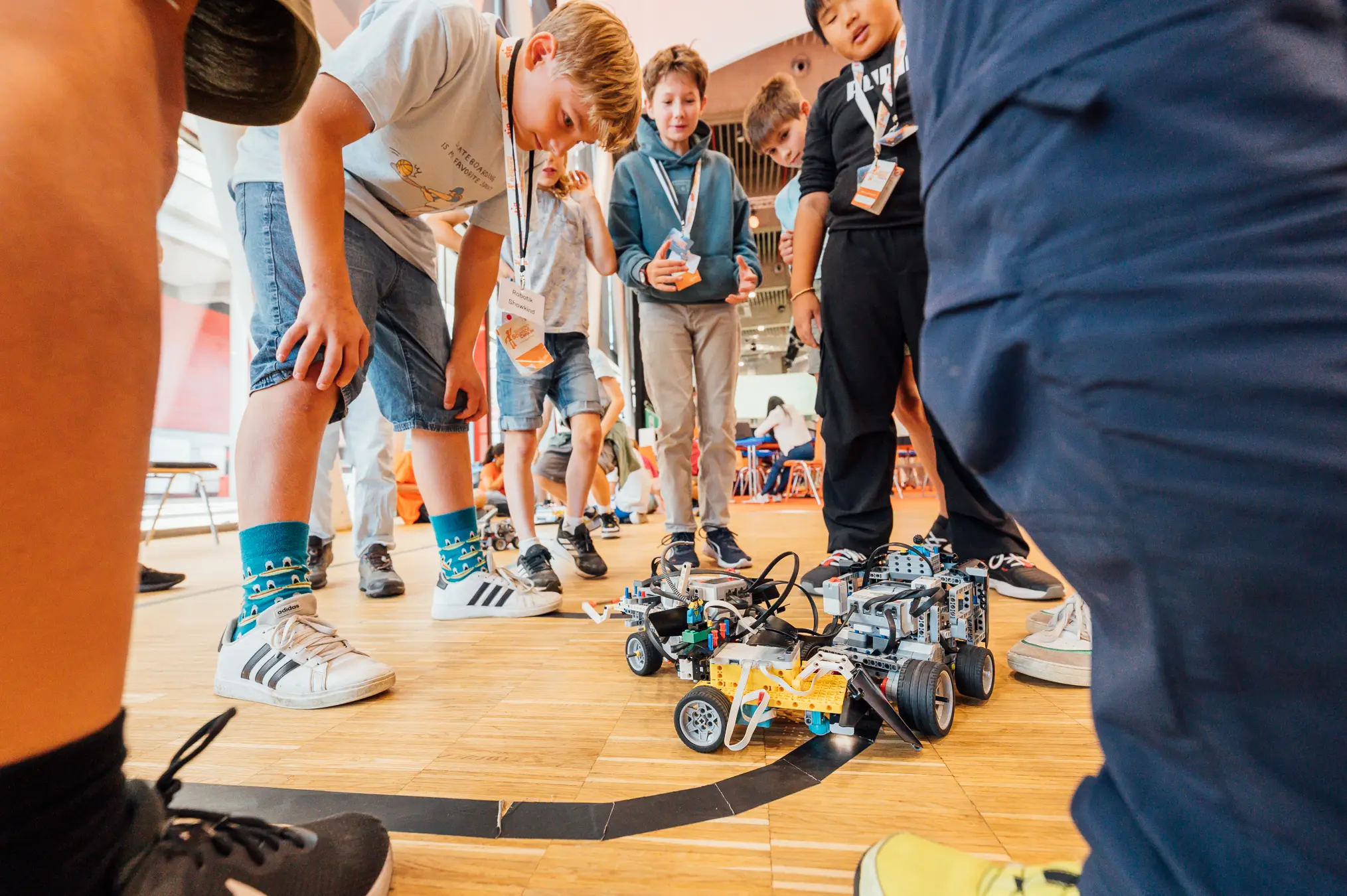 Kinder stehen im Kreis um selbstgebaute Lego-Roboter, die auf einer Linie am Boden fahren
